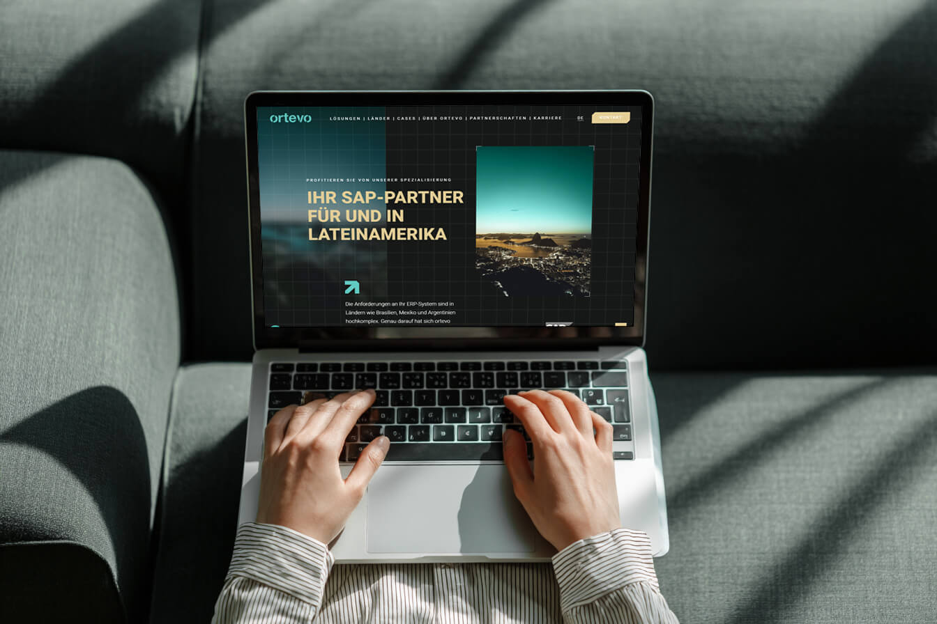 Eine Person tippt auf einem Laptop, auf dem eine Unternehmenswebsite mit dem deutschen Text Ihr SAP-Partner für und in Lateinamerika mit dem Schwerpunkt ki und automatisierung angezeigt wird, während sie auf einer grauen Couch sitzt und eine Stadtlandschaft im Blick hat. - NINE brackets Digitalagentur München