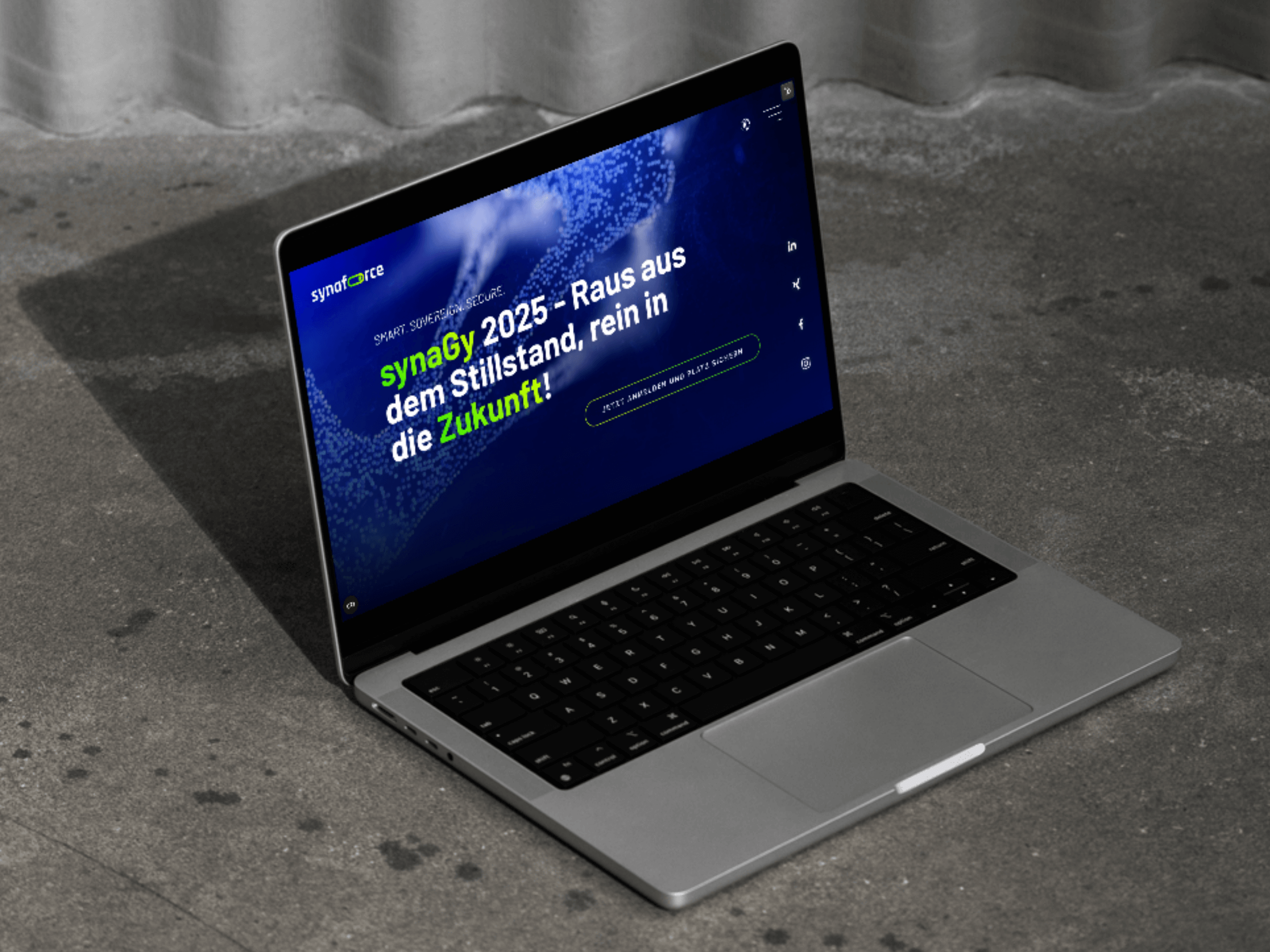 Ein Laptop steht auf einem Betonboden und zeigt eine Unternehmenswebsite mit der Überschrift: synaGY 2025 - Raus aus dem Stillstand, rein in die Zukunft! in deutscher Sprache, mit fetter grüner und weißer Schrift auf blauem Hintergrund. - NINE brackets Digitalagentur München