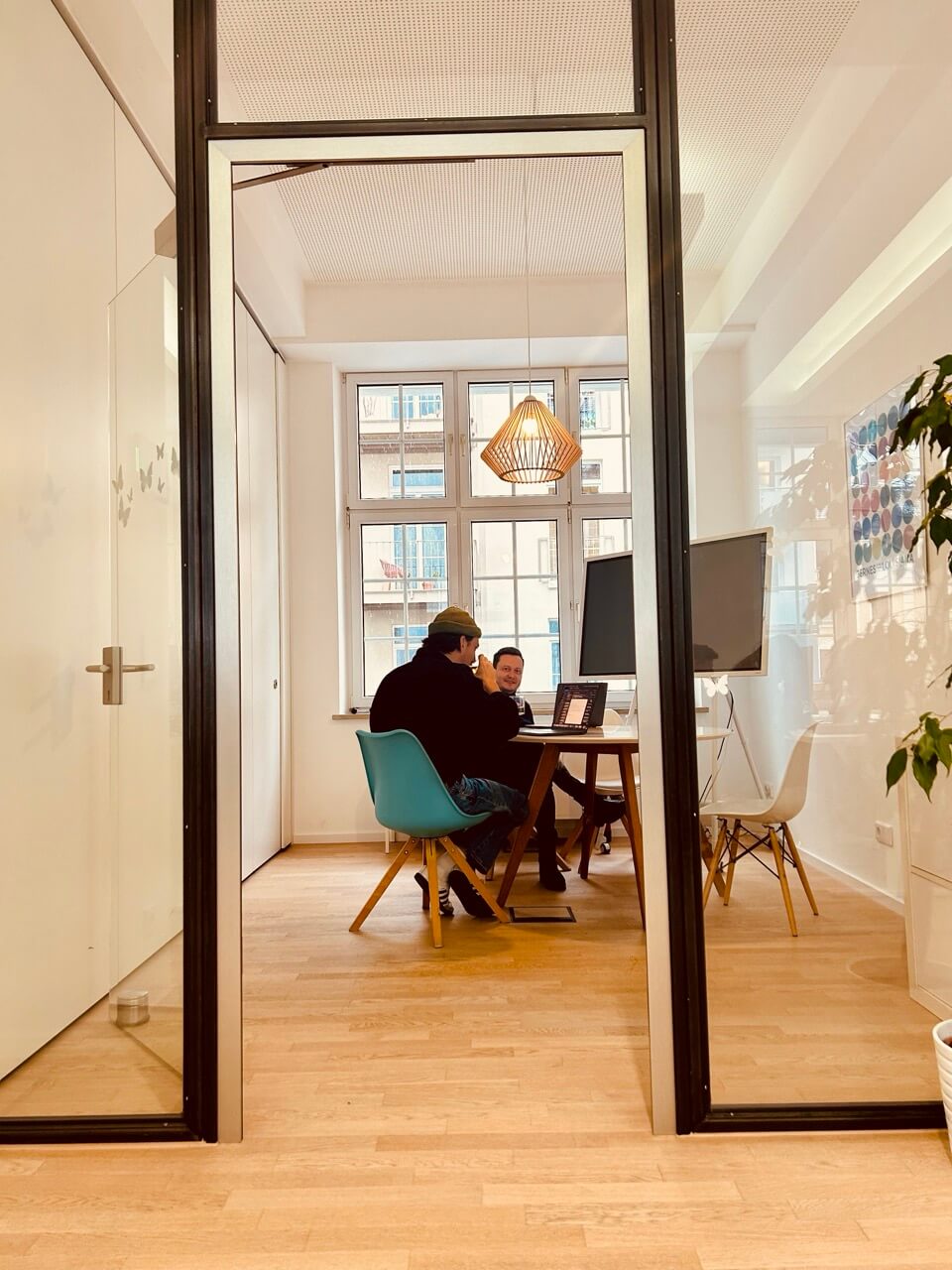 Zwei Personen sitzen an einem Tisch in einem modernen, hellen Büro der Digitalagentur München und arbeiten an Laptops. Große Fenster, Holzböden, Pflanzen und ein Whiteboard verschönern den Raum, in dem es um KI und Automatisierung geht. - NINE brackets Digitalagentur München