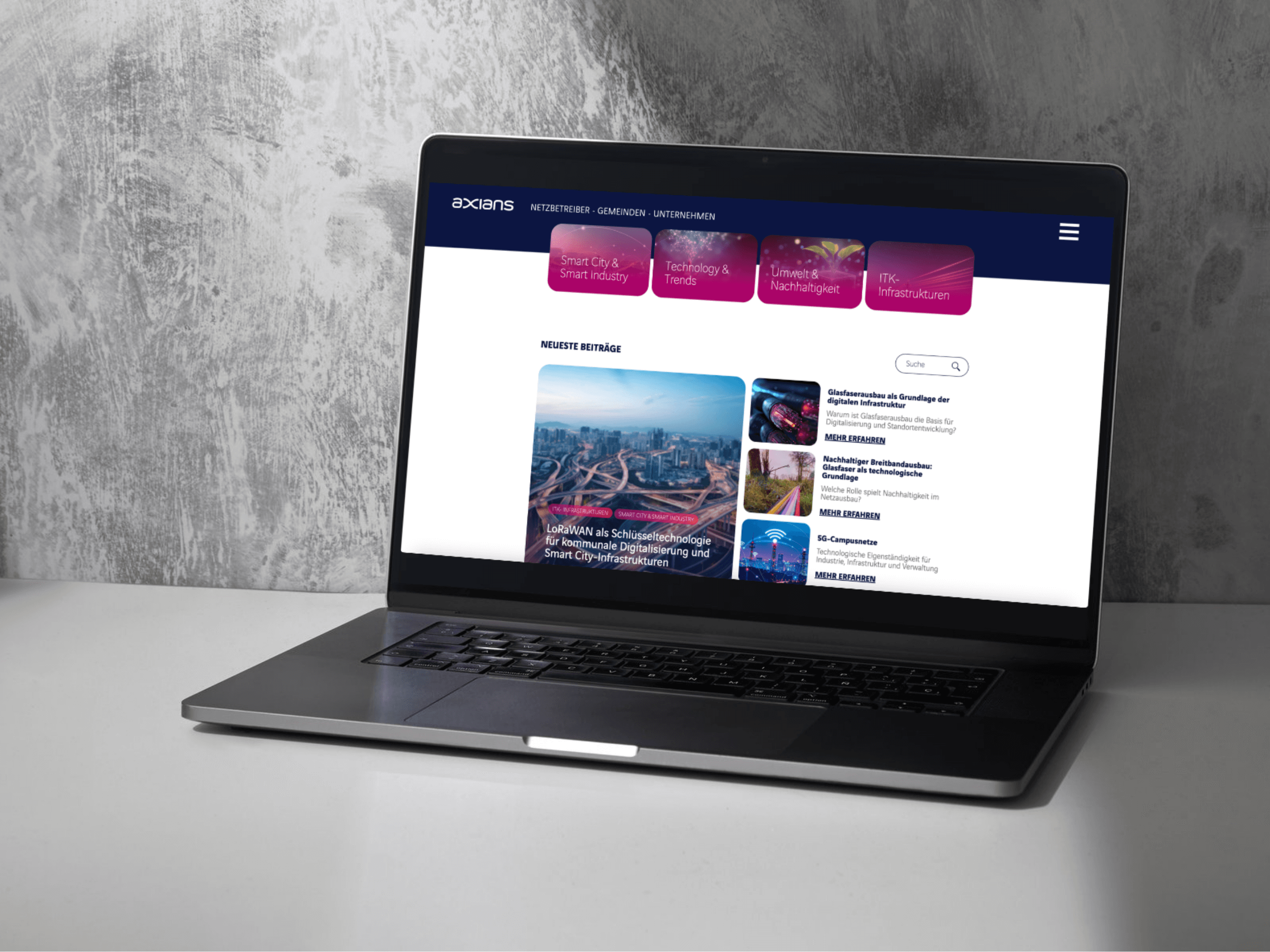 Ein Laptop auf einem grauen Schreibtisch zeigt eine Unternehmenswebsite mit einer blau-weißen Oberfläche, die Artikel und bunte Navigationsreiter am oberen Rand enthält. Das Setting deutet auf eine Digitalagentur München mit den Schwerpunkten KI und Automatisierung hin. Eine strukturierte graue Wand bildet den Hintergrund. - NINE brackets Digitalagentur München